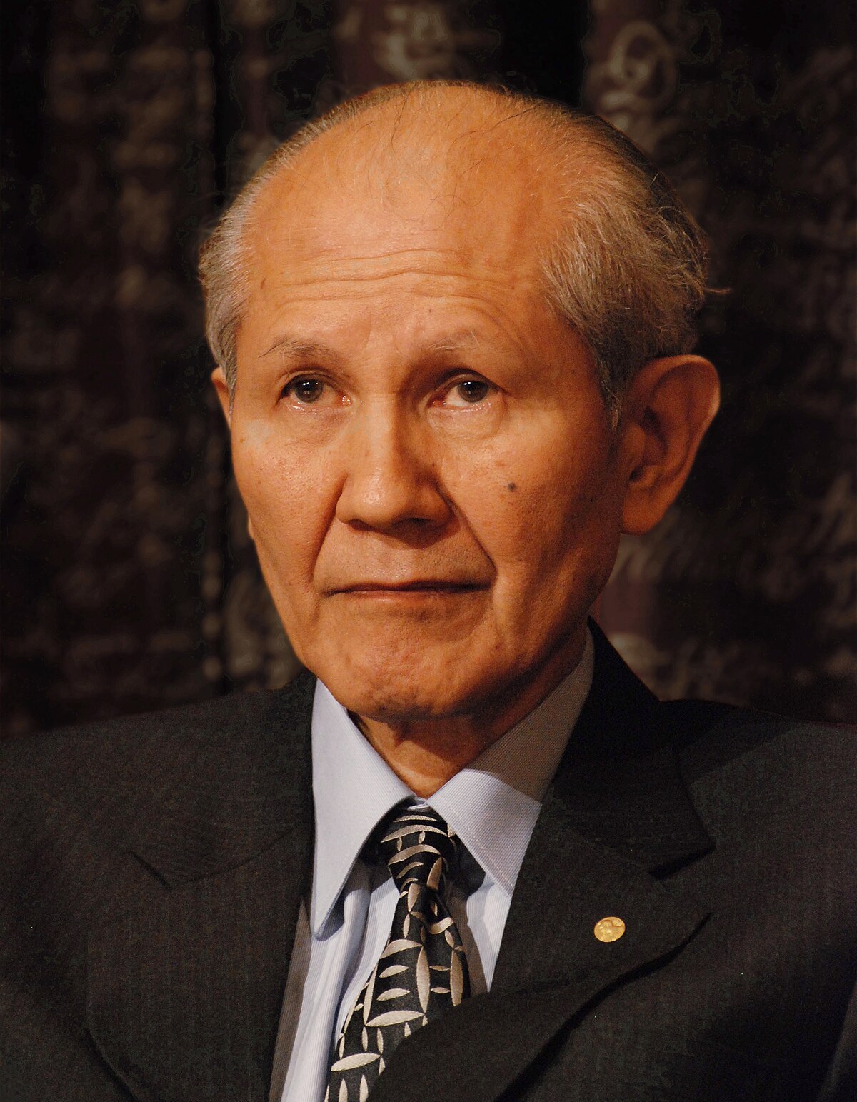 Portrait of Osamu Shimomura, a Nobel Prize laureate, wearing a dark suit and a patterned tie. He is looking directly at the viewer.