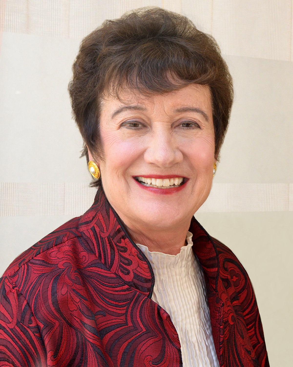 Portrait of Christina Maslach smiling, wearing a red and black patterned jacket over a white top, and gold earrings.