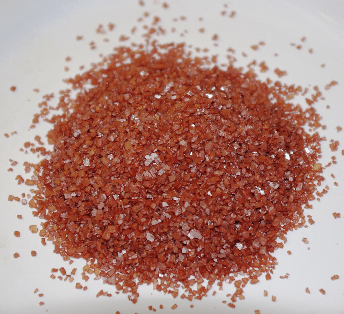 A close-up shot of a pile of reddish-brown alaea salt crystals on a white surface, showing individual grains with a slightly reflective quality.