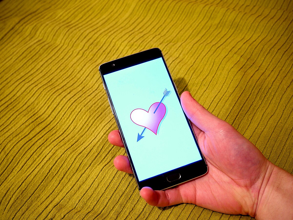 A hand holding a smartphone displaying a dating app illustration with a pink heart pierced by a blue arrow on its screen. The phone is placed on a textured yellow-green fabric.