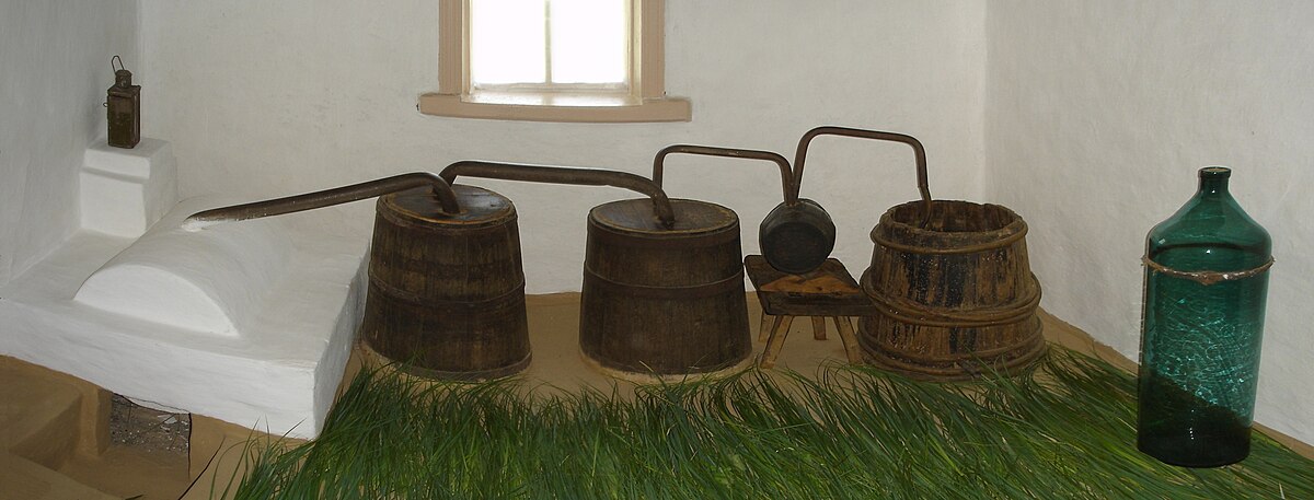 An old Ukrainian vodka still displayed indoors, featuring three wooden barrels connected by metal pipes, suggesting a traditional distillation process. The setup is placed on a bed of straw, with a window and a bottle visible in the background.
