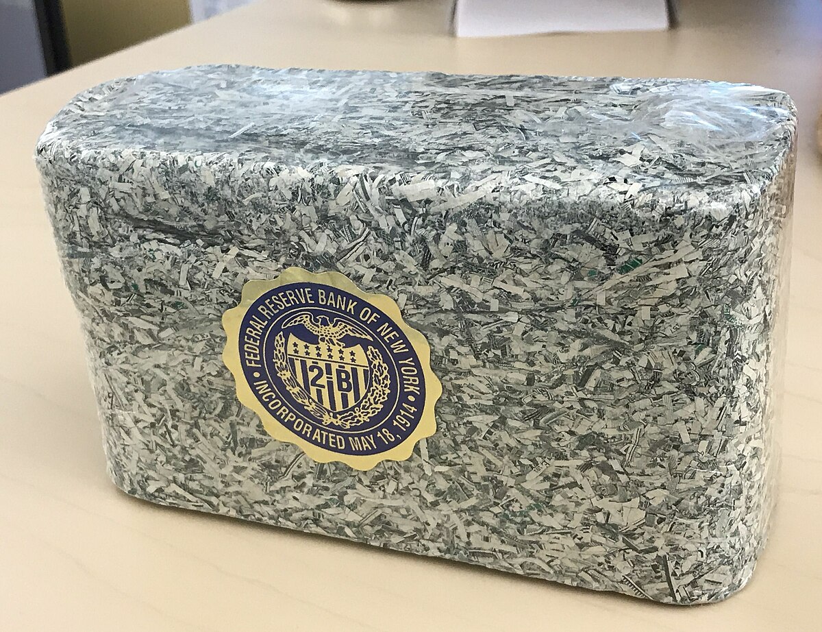 A rectangular block of shredded US Dollar notes, wrapped in clear plastic, with a circular seal from the Federal Reserve Bank of New York. The seal reads "FEDERAL RESERVE BANK OF NEW YORK" and "INCORPORATED MAY 18, 1914" around an eagle emblem.