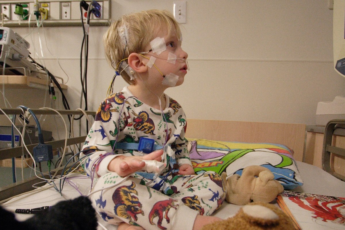 A young child, a pediatric polysomnography patient, is shown sitting up in a hospital bed with numerous wires and sensors attached to their head, face, and body. The child is wearing patterned pajamas and looking to the right, with medical equipment visible in the background.