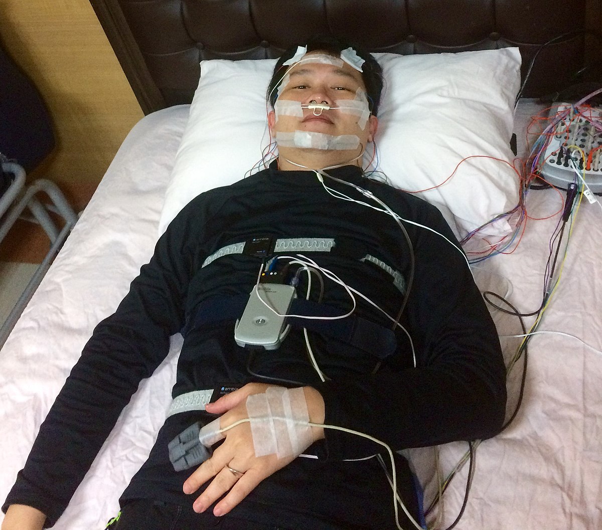 A patient is shown lying in bed with polysomnography equipment attached for an overnight sleep study. Sensors are placed on the head, face, chest, and finger, connected to a recording device beside the bed.