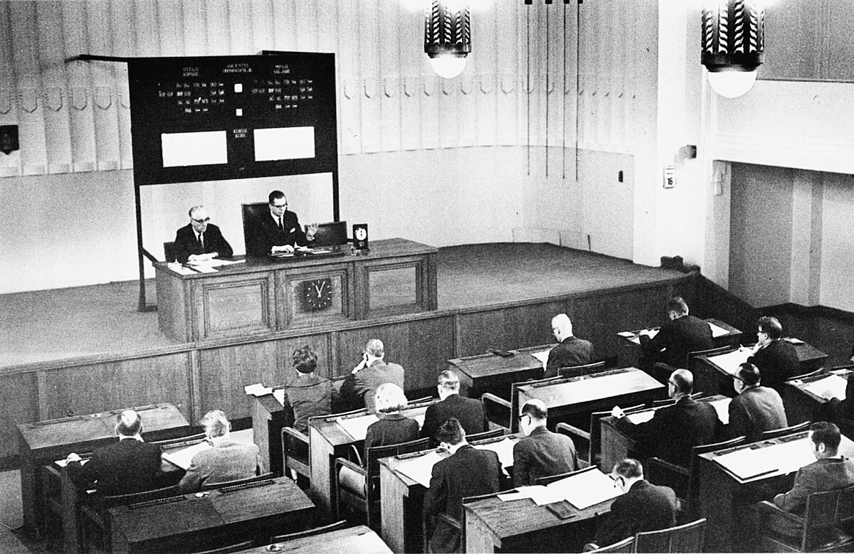 A black and white photograph of the Helsinki Stock Exchange in 1965, showing a trading room with two men at a central desk on a raised platform, facing a large display board. Several other men are seated at desks in the foreground, observing the proceedings.
