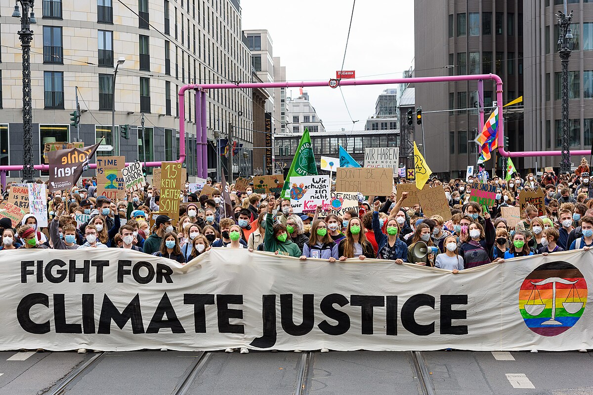 A large crowd of people, including Greta Thunberg and Luisa Neubauer, are gathered at a Fridays for Future demonstration in Berlin in September 2021, holding signs and a large banner that reads "FIGHT FOR CLIMATE JUSTICE." Many participants are wearing face masks.