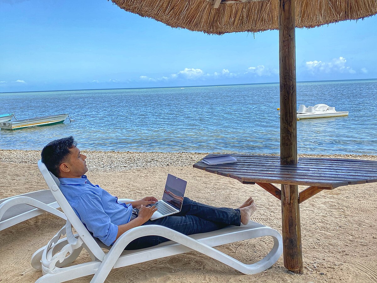 A man working on a laptop while relaxing on a lounge chair under a straw umbrella on a sandy beach. The calm blue ocean stretches into the horizon, with small boats visible in the distance, suggesting a remote work or digital nomad lifestyle.