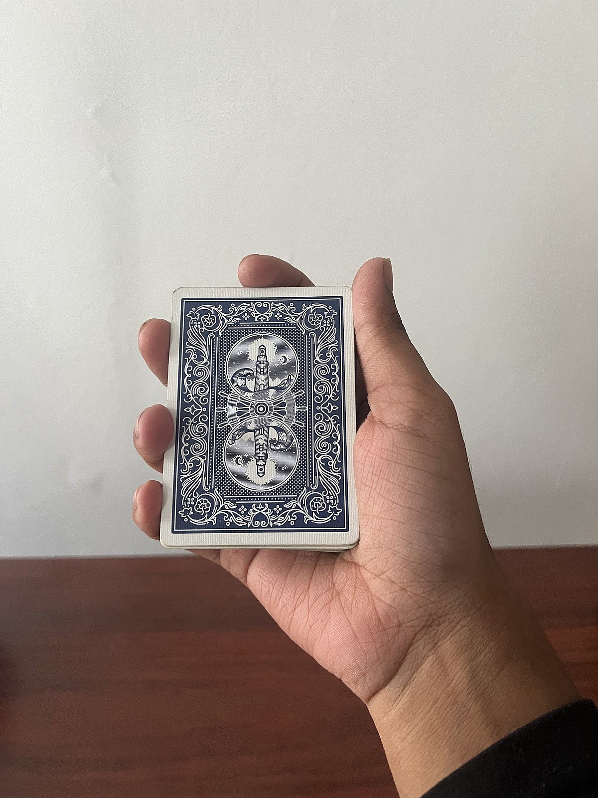 A hand holds a deck of playing cards in a mechanic's grip, with the blue-backed cards facing the viewer. The card design features a symmetrical pattern of swords, shields, and moons.
