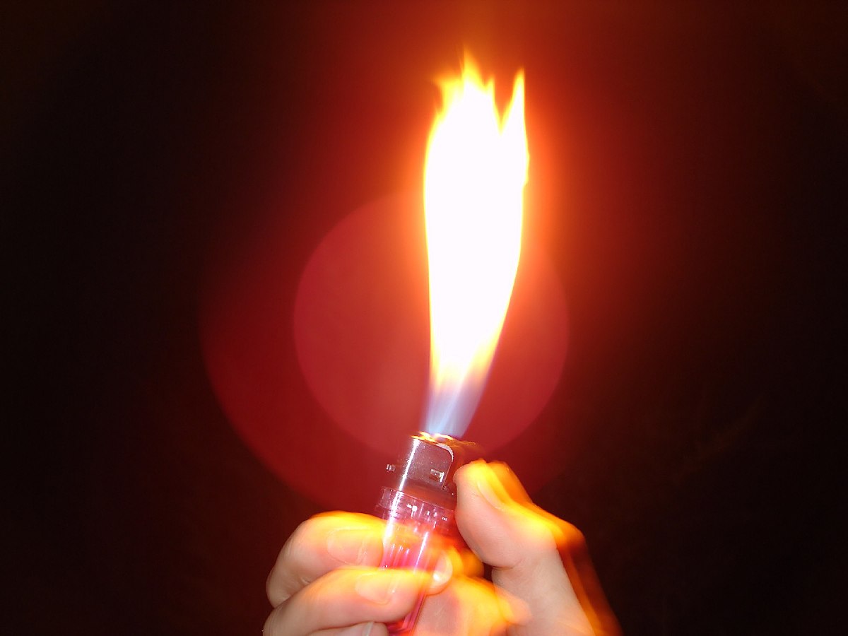 A hand holding a lit lighter, producing a bright flame against a dark background with a red glow. The image captures the motion blur of the hand and flame.