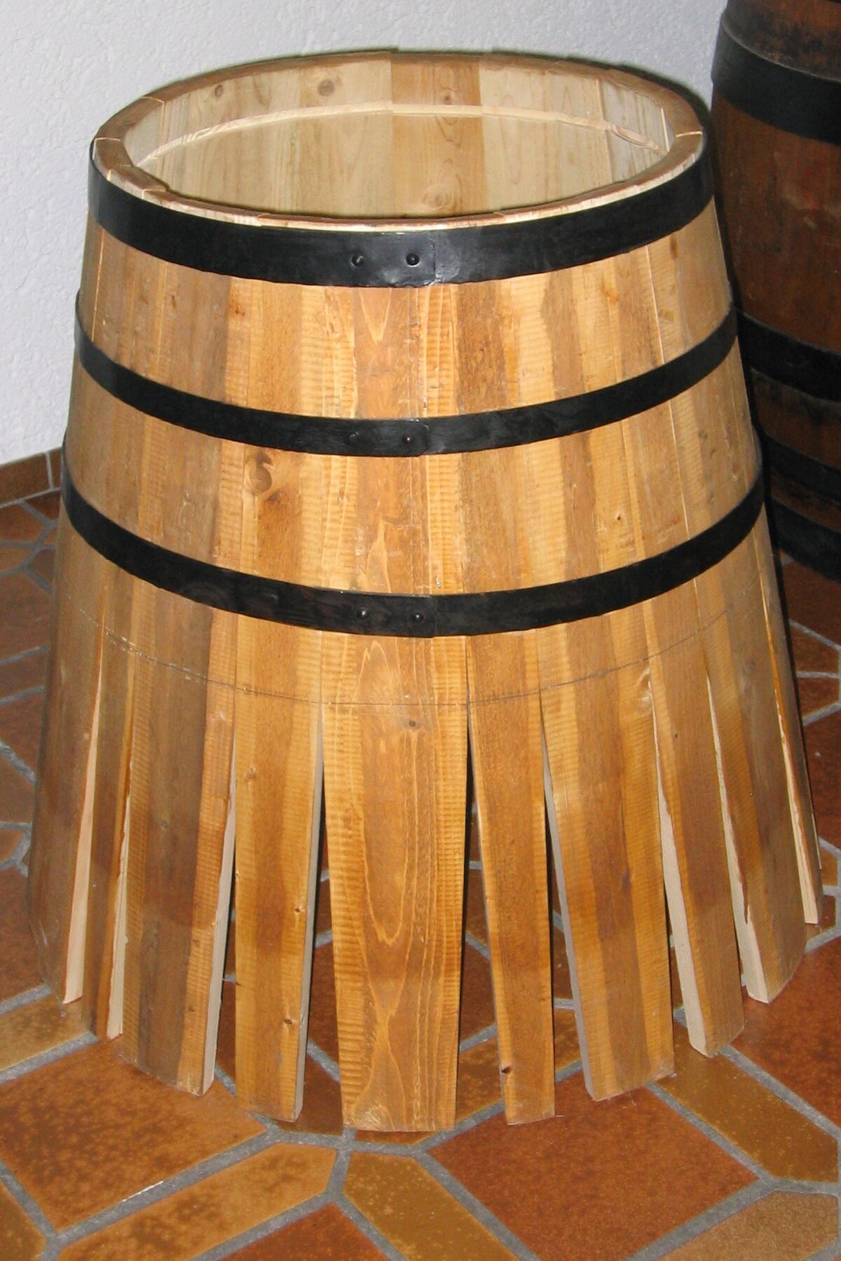 A wooden barrel midway through construction, with its staves held together by black metal hoops, but the bottom and top are not yet fully assembled.