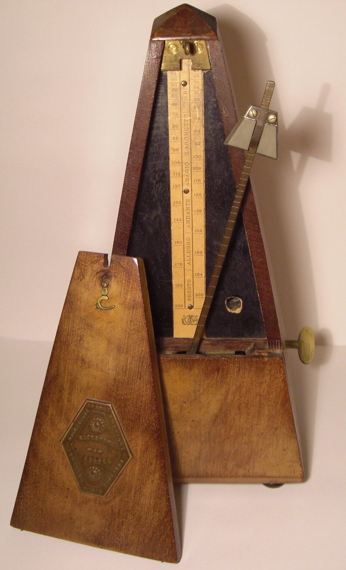 A Maelzel's Metronome, a pyramid-shaped wooden device, is shown with its front cover removed, revealing a pendulum and a scale marked with tempo indications. The scale includes numbers from 40 to 208 and Italian tempo terms such as "LARGO," "ADAGIO," "ANDANTE," "ALLEGRO," and "PRESTO."