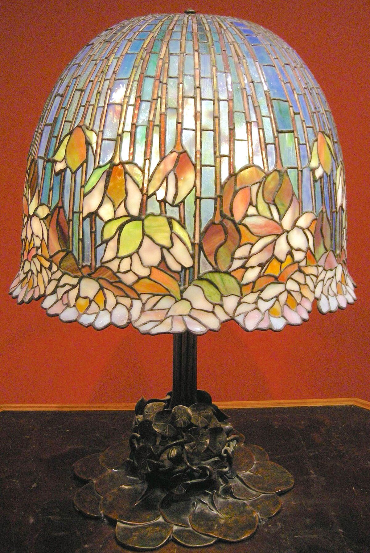 A Louis Comfort Tiffany table lamp, Pomb Lily model, from approximately 1900-1910, featuring a domed stained-glass shade with a pattern of water lilies in various colors. The base of the lamp is made of bronze with a lily pad motif.