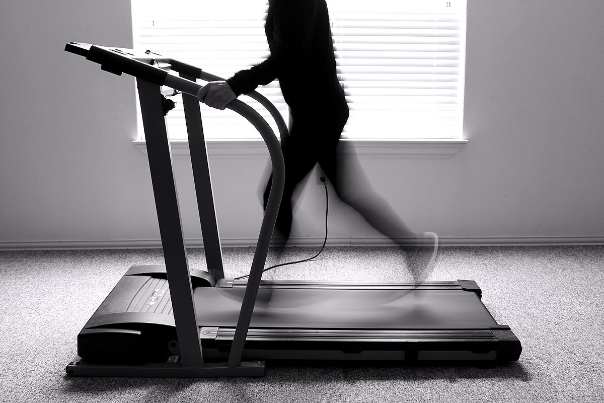 A black and white photograph of a person running on a treadmill, with their motion blurred to convey movement, suggesting aerobic exercise.