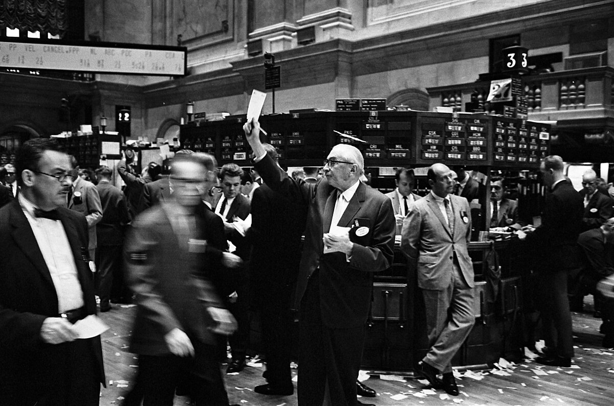 The NYSE's stock exchange traders floor in 1963 shows numerous traders in suits, with one man in the foreground raising a piece of paper. The floor is active, with some paper scattered, and an overhead display board is visible.