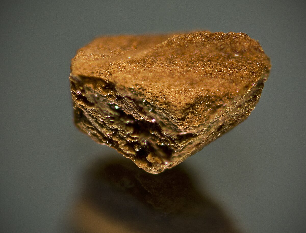 A close-up view of a single granule of Nescafé instant coffee, showing its irregular shape and porous, crystalline texture. The granule is brown and appears to be floating above a reflective surface.