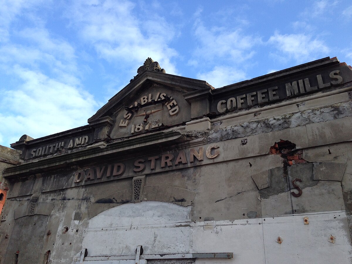 The facade of the David Strang Coffee Mills building, established in 1874, features weathered stone and brick with the embossed text "ESTABLISHED 1874" and "DAVID STRANG COFFEE MILLS." The building shows signs of age and disrepair under a blue sky.