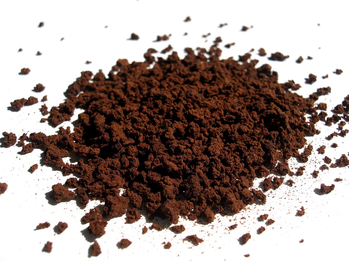 A pile of dark brown instant coffee powder is scattered on a white surface, showing individual granules and finer particles. The coffee appears dry and granular, with some variation in particle size.