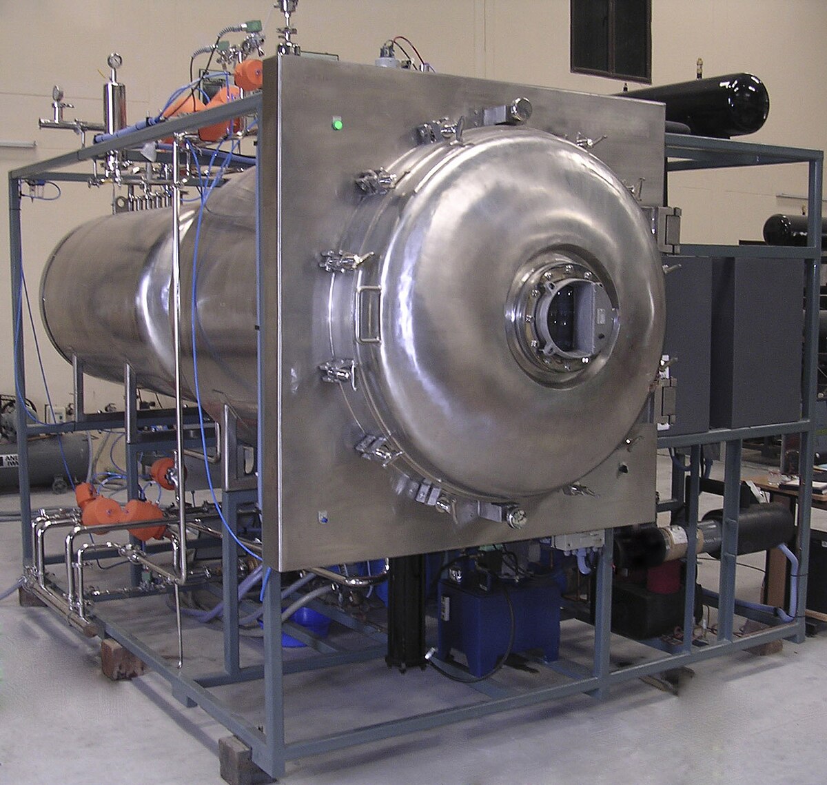 A large industrial freeze dryer, a complex piece of machinery with a cylindrical vacuum chamber and numerous pipes and controls, is shown in a production setting. The main chamber is made of stainless steel with multiple clamps and a viewing port.