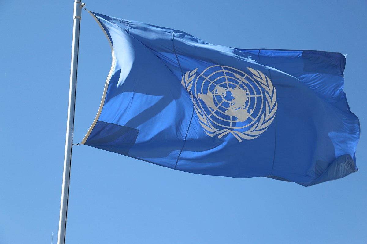 The blue flag of the United Nations, featuring a white emblem of the world map flanked by olive branches, waves against a clear blue sky. The flag is attached to a flagpole on the left side of the image.
