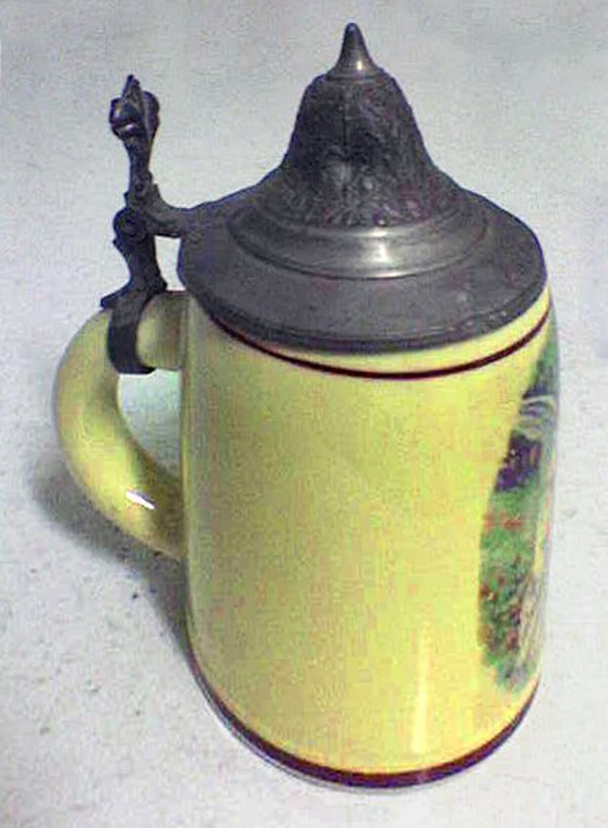 A ceramic tankard with a yellow body and a pewter lid is shown from an angled perspective. The tankard features a colorful landscape illustration on its side.