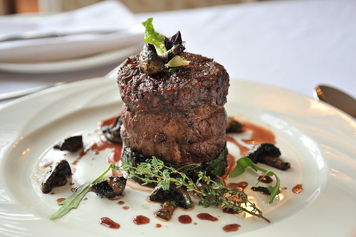 A beef fillet steak dinner is served with mushrooms, garnished with herbs, and drizzled with sauce on a white plate.