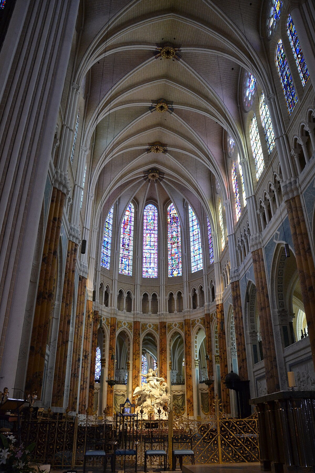 Interior view of Chartres Cathedral, highlighting its soaring vaulted ceilings, tall columns, and numerous stained-glass windows that fill the space with colorful light. A large white sculptural piece is visible in the center of the image.