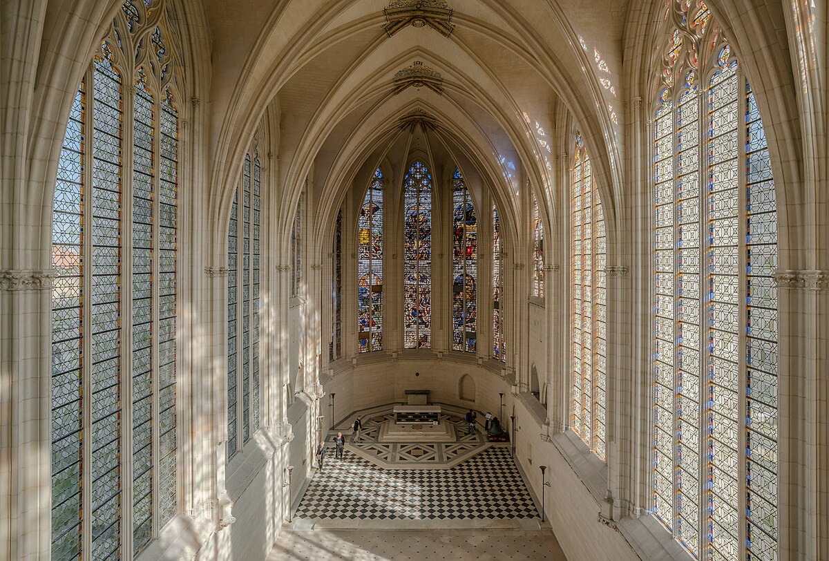 An aerial view of the interior of Sainte-Chapelle in Vincennes, showing the high vaulted ceilings and numerous tall stained glass windows. The altar is visible at the far end, with a black and white checkered floor leading up to it.