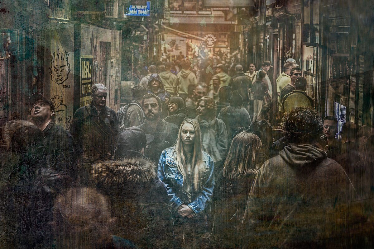 An artistic depiction titled "Loneliness in a Crowd," showing a young woman standing alone and looking directly at the viewer, surrounded by a blurred and indistinct crowd of people in an urban setting. The image conveys a sense of isolation despite being in a populated area.
