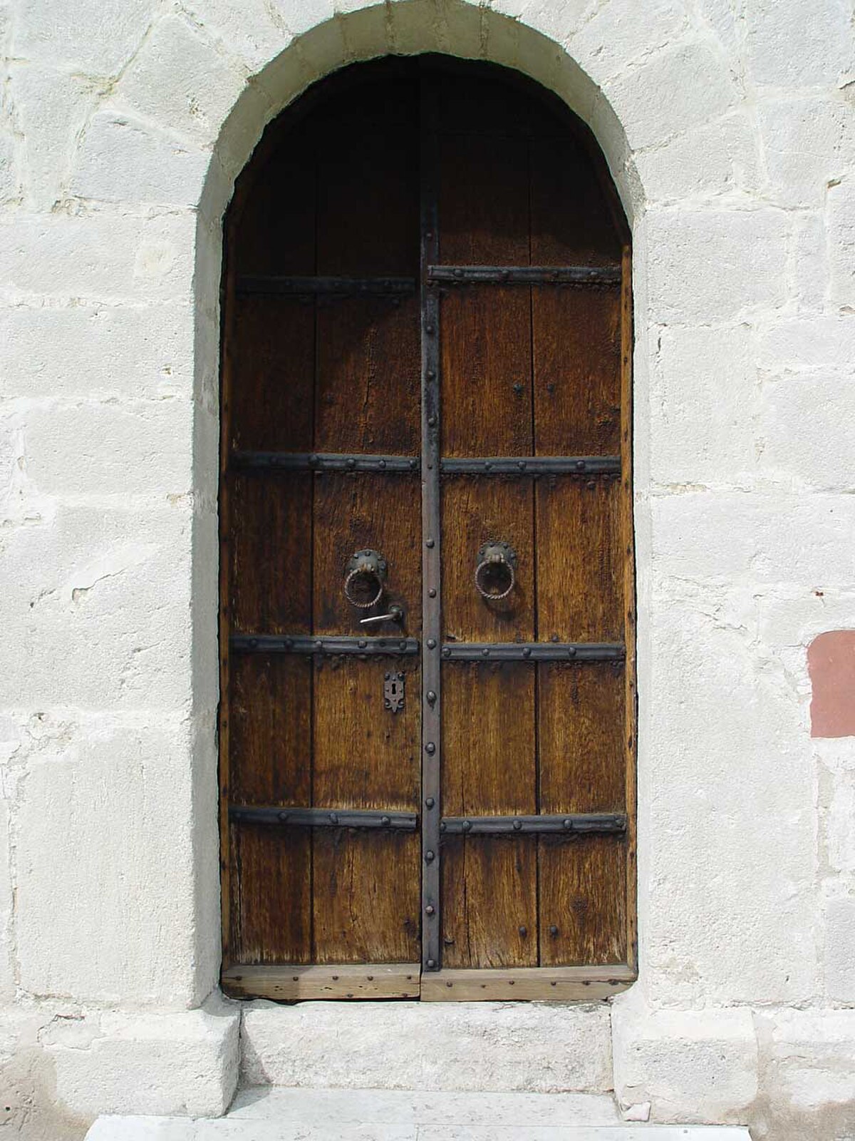 An arched wooden door with metal bands and two ring handles, set within a stone archway. The door is closed, suggesting a hidden interior.