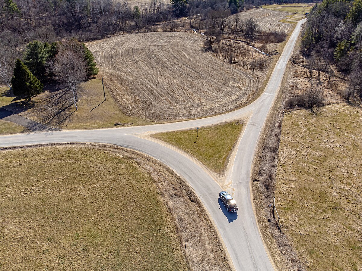 Aerial view of a Y-junction, or "fork in the road," in a rural setting, with two paved roads diverging. The surrounding landscape consists of bare trees, brown fields, and patches of green grass, with a single car visible on one of the roads.