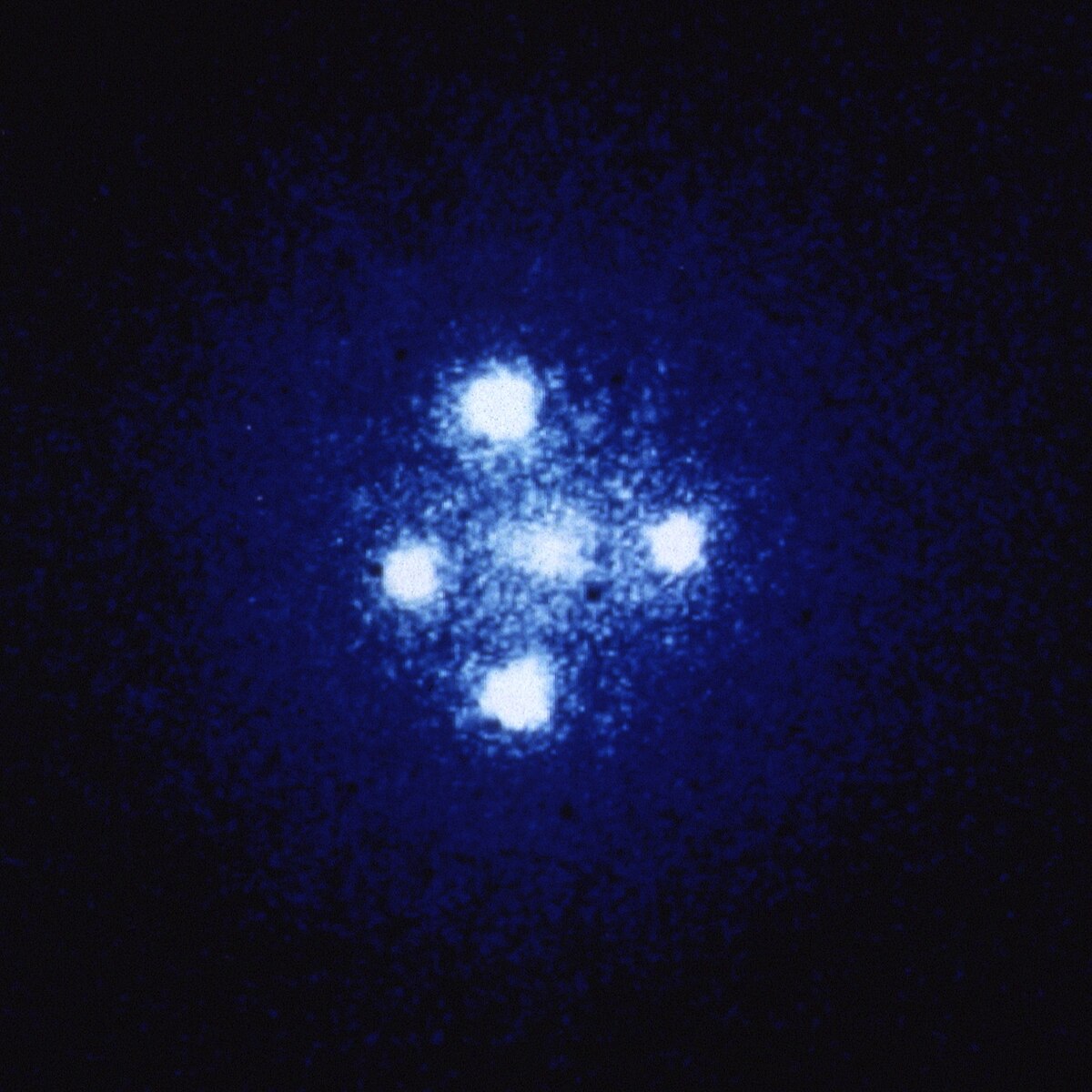 An image of the Einstein Cross, showing four bright, blue-white points of light arranged in a cross pattern around a central, fainter object. These four images are of the same astronomical object, gravitationally lensed by a foreground galaxy.