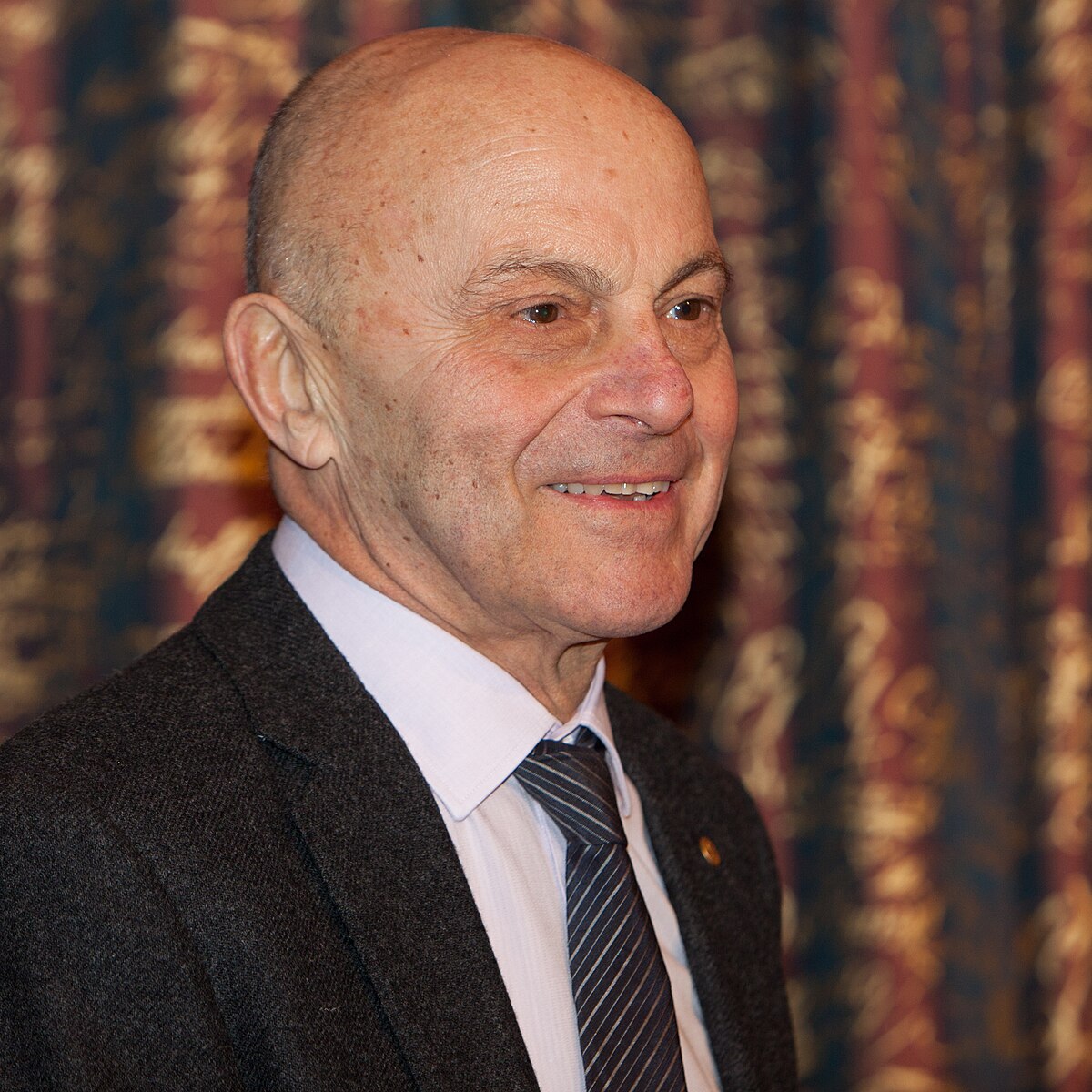 A portrait of Eugene Fama, a Nobel laureate in economics, smiling and looking to the right. He is wearing a dark suit and a striped tie.