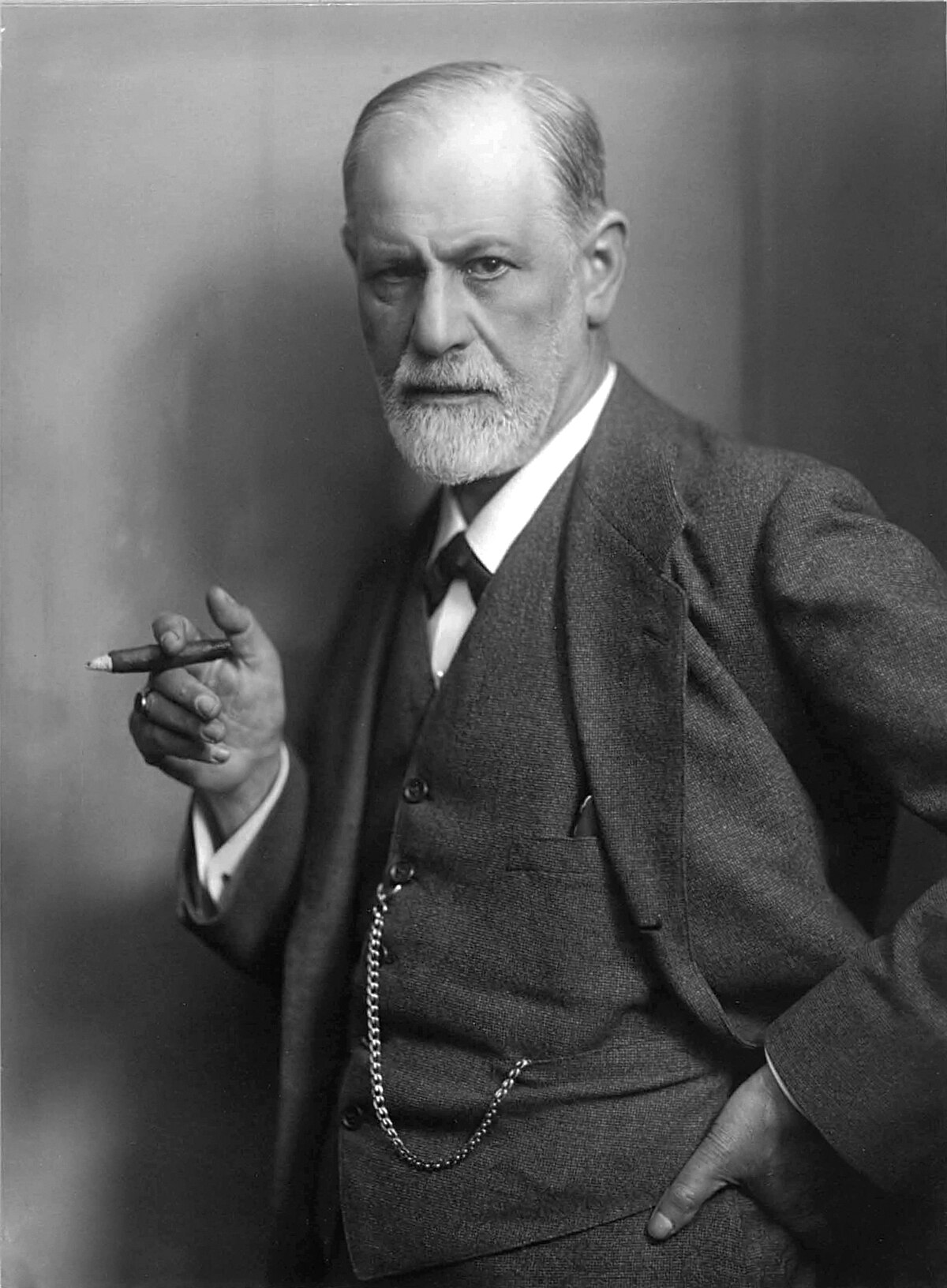 A black and white portrait of Sigmund Freud, taken around 1921 by Max Halberstadt, shows him holding a cigar. He is dressed in a suit with a waistcoat and a bow tie, looking directly at the viewer.