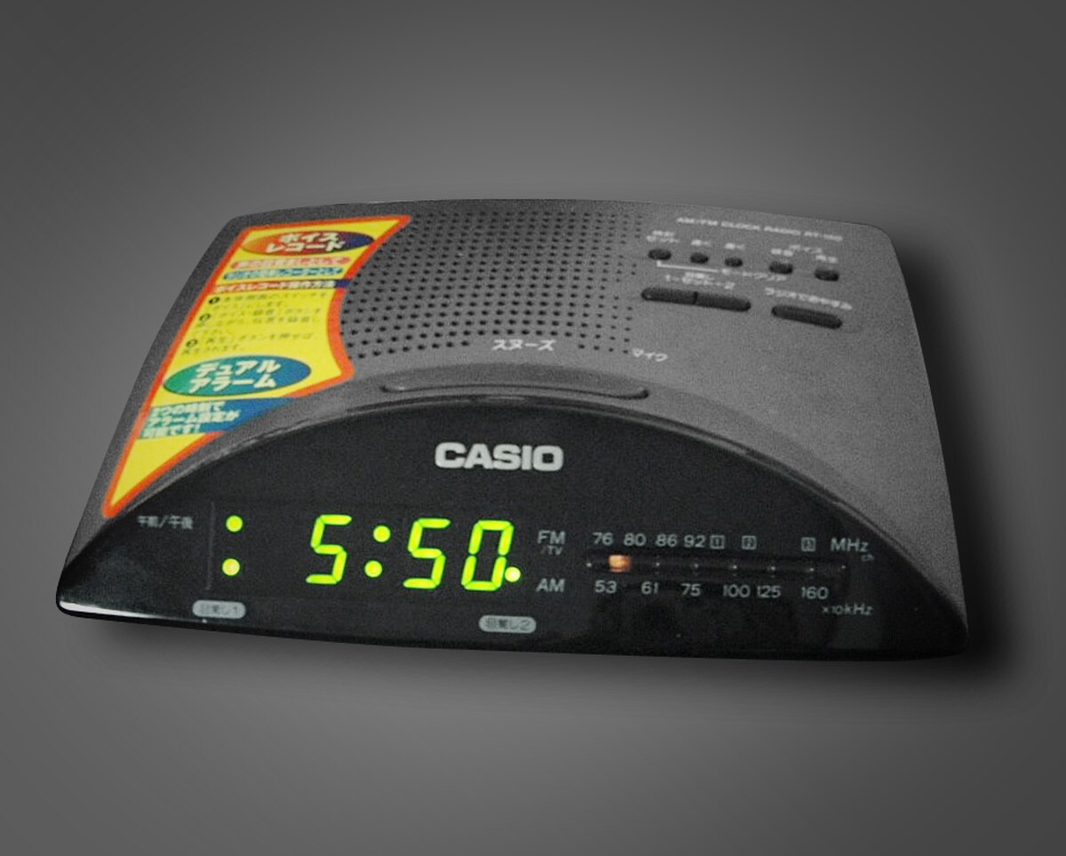 A Casio clock radio is shown with a digital display indicating the time "5:50" and an analog tuning dial for AM and FM radio frequencies. The device combines a broadcast radio receiver with an alarm clock function, allowing users to wake up to radio audio.