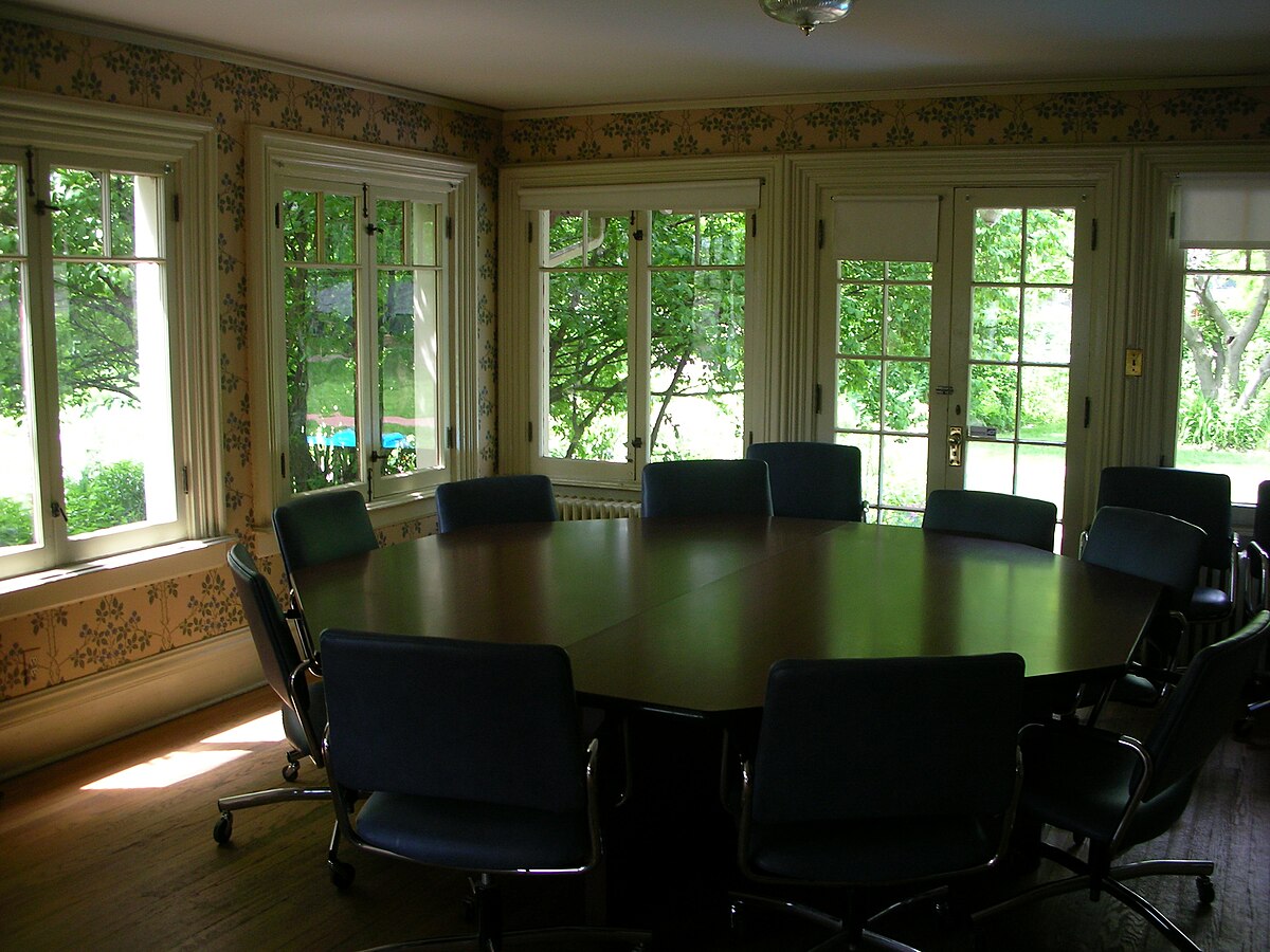 A classroom or meeting room featuring a large octagonal table surrounded by chairs, with multiple windows looking out onto a green outdoor scene. The room has patterned wallpaper and a chandelier.