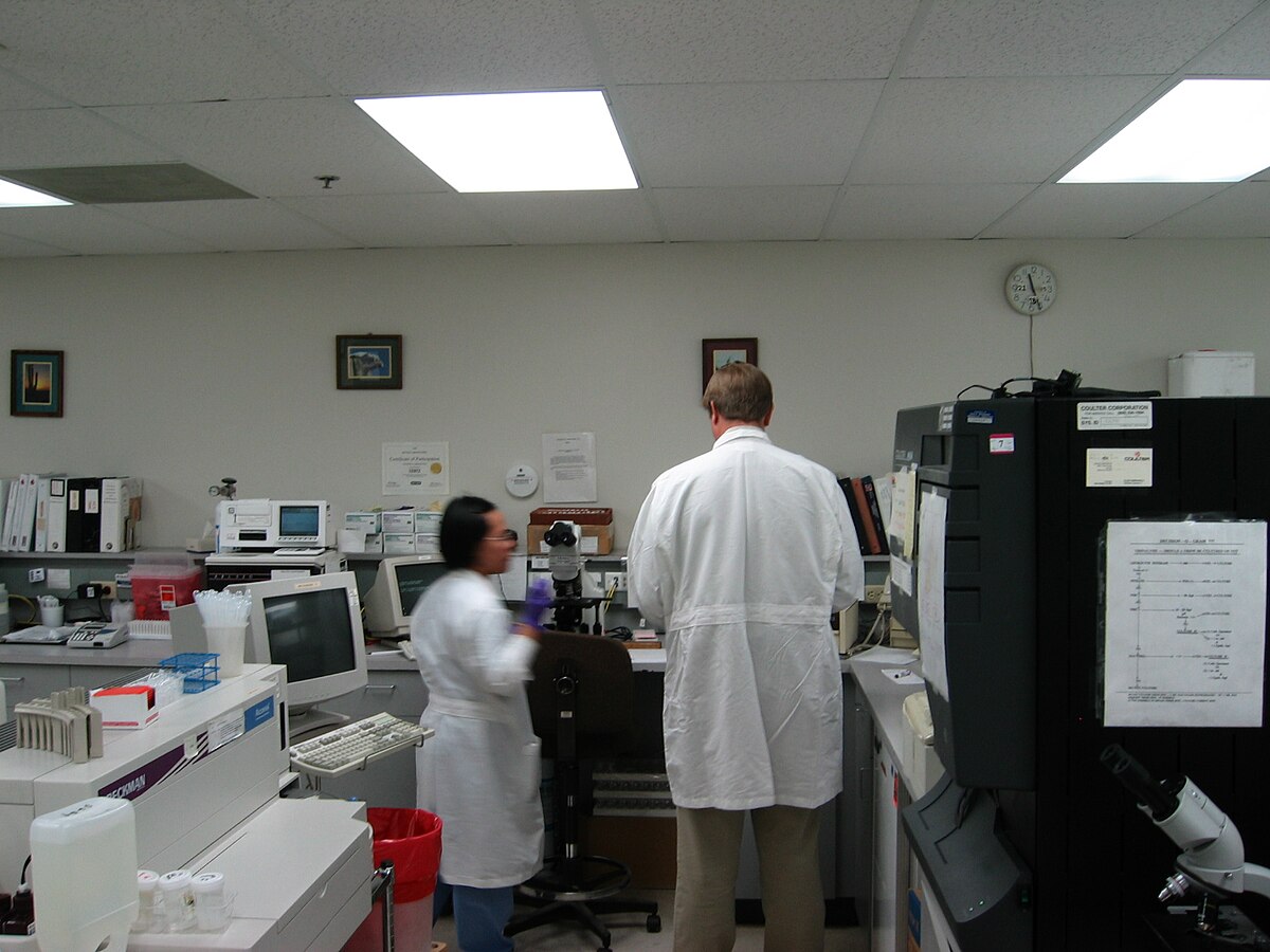 Clinical laboratory in a hospital setting with two technologists, one facing away in a white lab coat and another facing left, wearing a blue shirt and lab coat. The lab features various equipment, including computers, microscopes, and automated analyzers, with shelves of binders and supplies in the background.