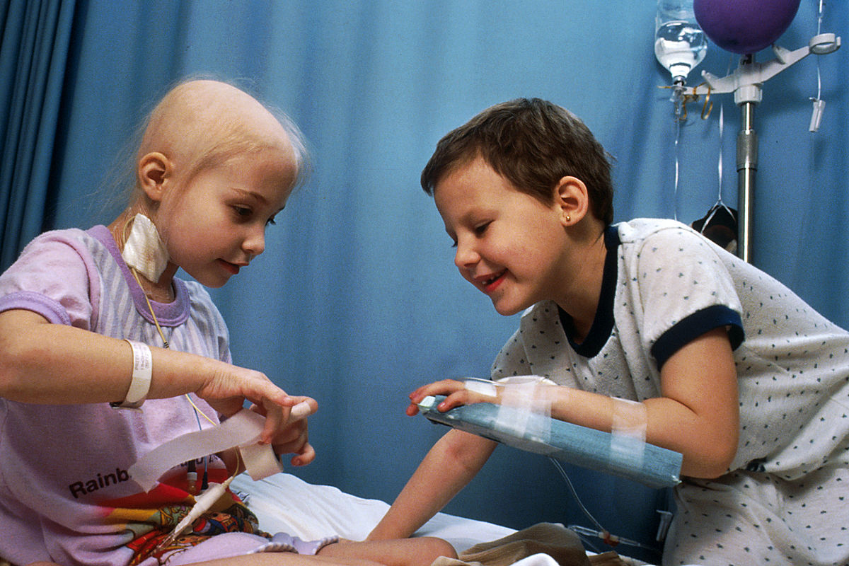 Two pediatric patients, a girl with a bald head and a boy, are shown in a hospital setting, both with intravenous catheters in their arms, suggesting they are receiving chemotherapy. They are smiling and interacting, with medical equipment visible in the background.