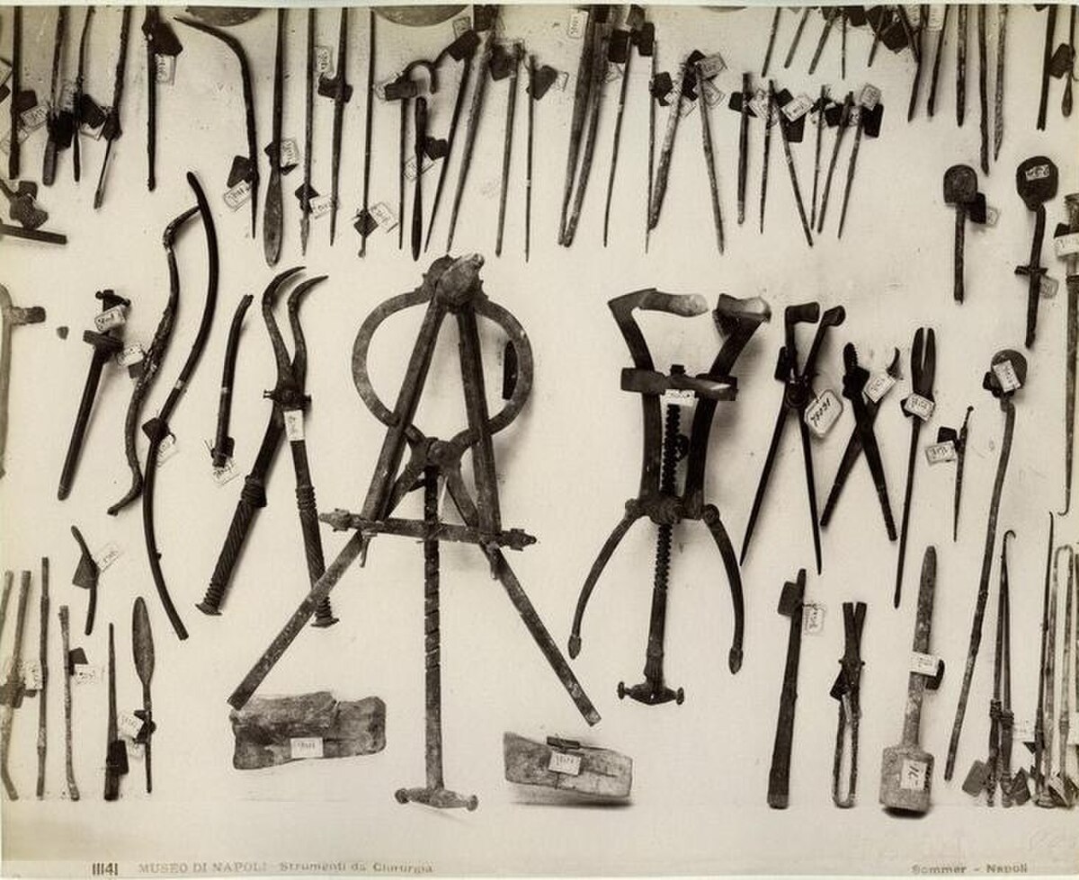 A collection of ancient Roman surgical instruments, including various forceps, hooks, scalpels, and a large tripod-like device, displayed against a dark background. The image is a historical photograph by Giorgio Sommer, labeled "Museo di Napoli, Strumenti di chirurgia."