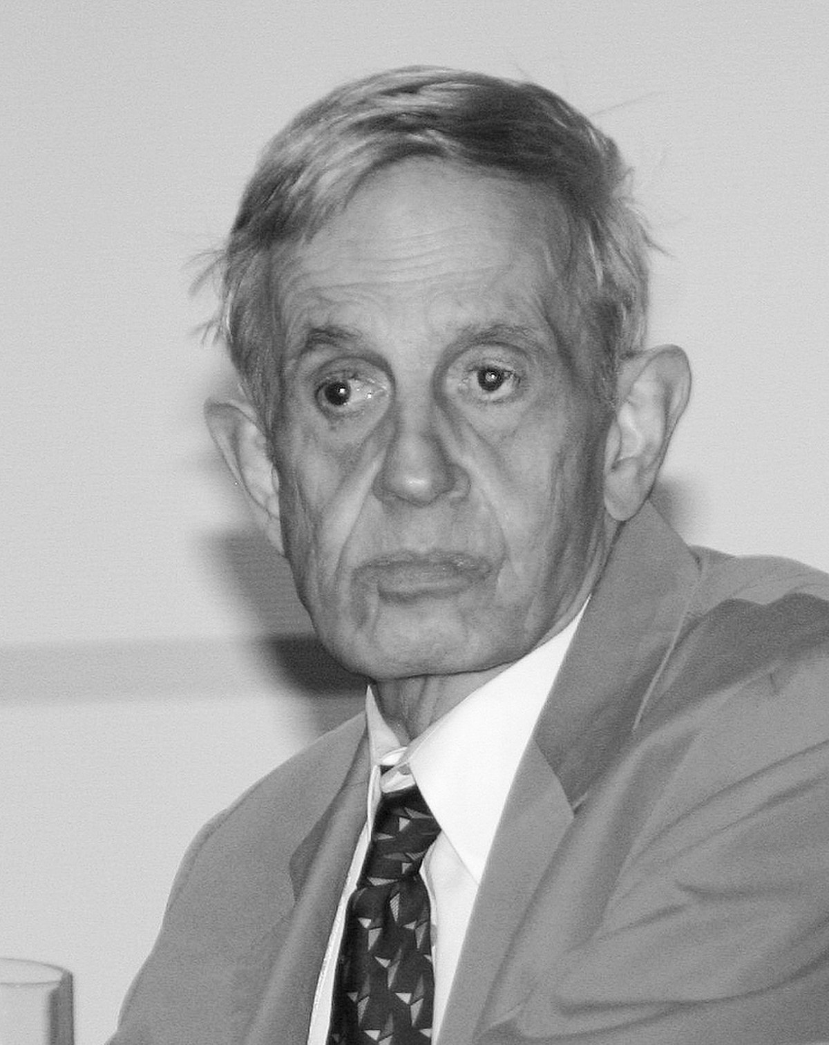 Black and white portrait of John Nash, an older man with a pensive expression, wearing a suit and a patterned tie, looking to his right.
