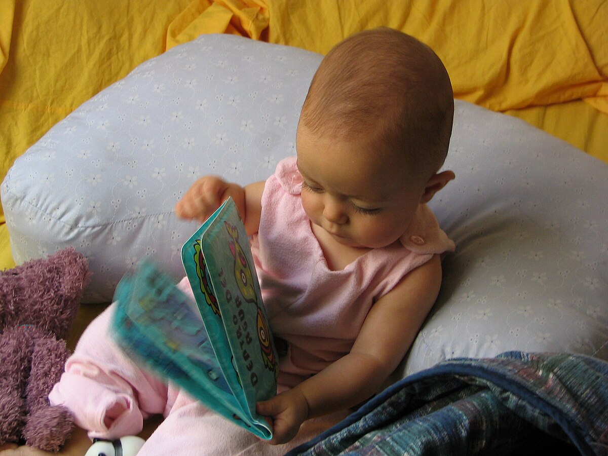 A baby in a pink outfit sitting on a pillow, looking at and interacting with a colorful board book. The baby is holding the book open with both hands.