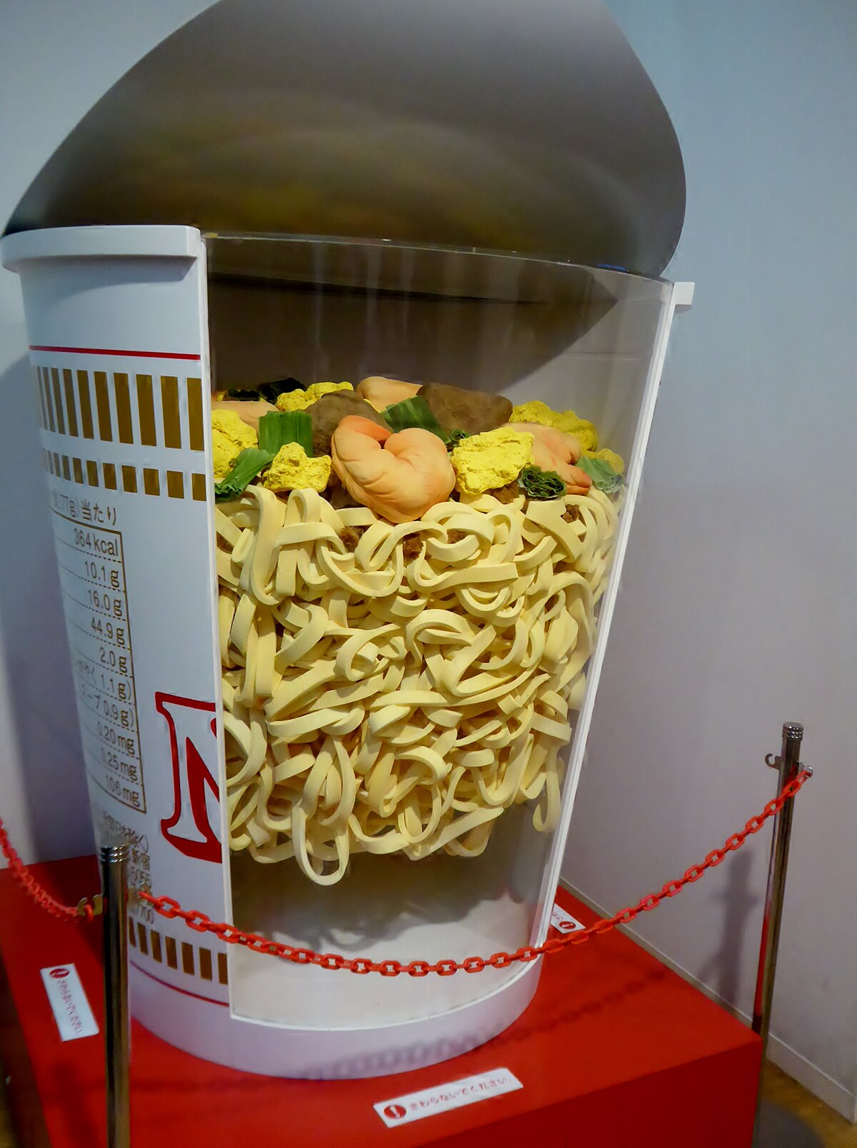 A cross-section model of a Cup Noodle shows the layered composition of instant noodles and various toppings inside a large white cup. Nutritional information in Japanese is visible on the side of the cup.
