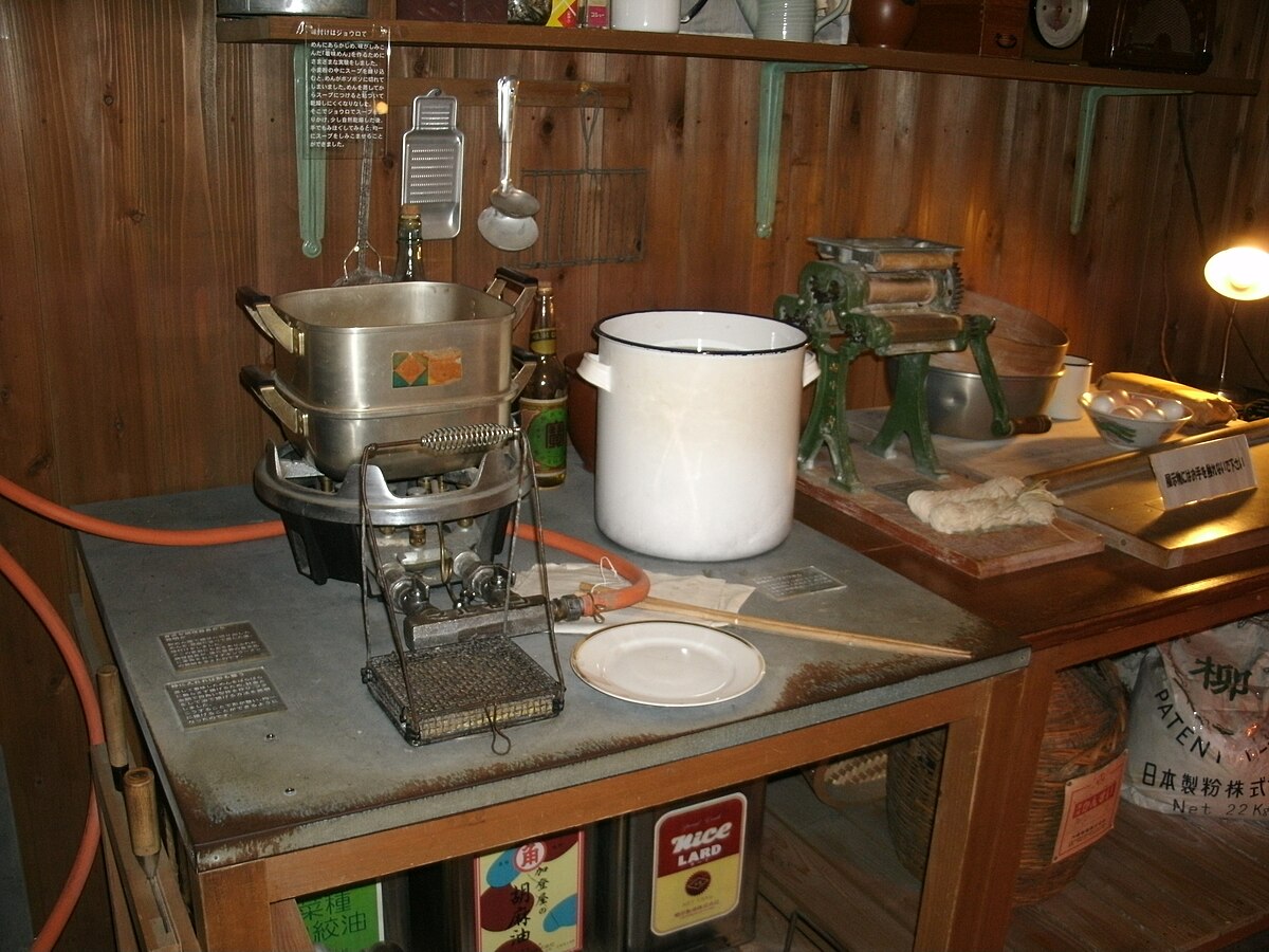 A recreation of Momofuku Ando's workshop, where instant noodles were created, features a vintage kitchen setup with a stovetop, pots, and a noodle press. Various cooking utensils hang on the wooden wall, and bags labeled "RICE LAND" and "日清製粉" (Nisshin Seifun) are visible on a shelf below the counter.
