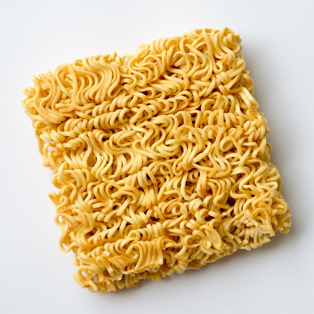 A rectangular block of Mama brand instant noodles, characterized by its wavy, dehydrated strands, is shown on a white background. The noodles are a light yellow color.