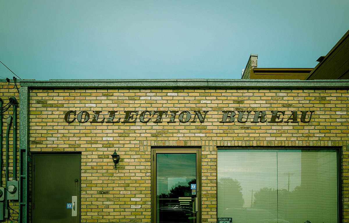 A brick building with the words "COLLECTION BUREAU" in raised letters above the entrance, located in Little Falls, Minnesota. The building has a glass door and a smaller solid door.