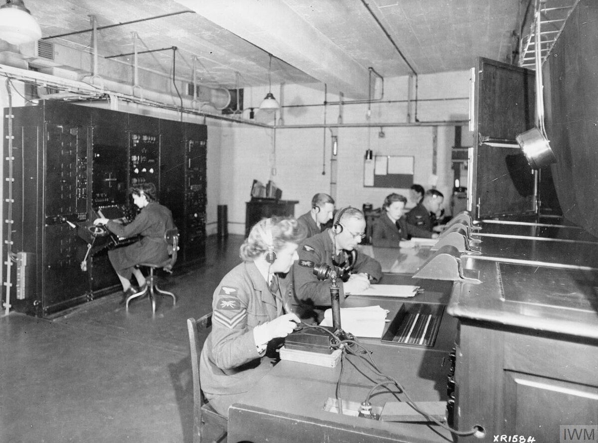 East Coast Chain Home radar operators, primarily women, are shown working at their stations in England. They are seated at consoles with headphones, monitoring equipment, and making notes, with large electronic cabinets visible in the background.