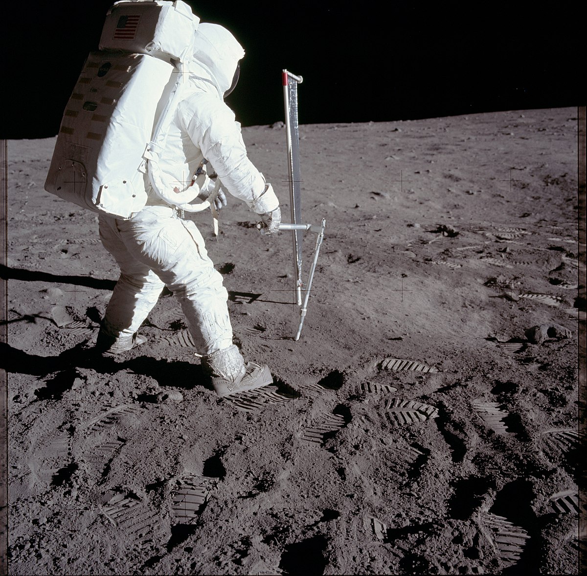 Astronaut Buzz Aldrin is depicted taking a core sample on the Moon during the Apollo 11 mission. His footprints are clearly visible in the lunar dust.