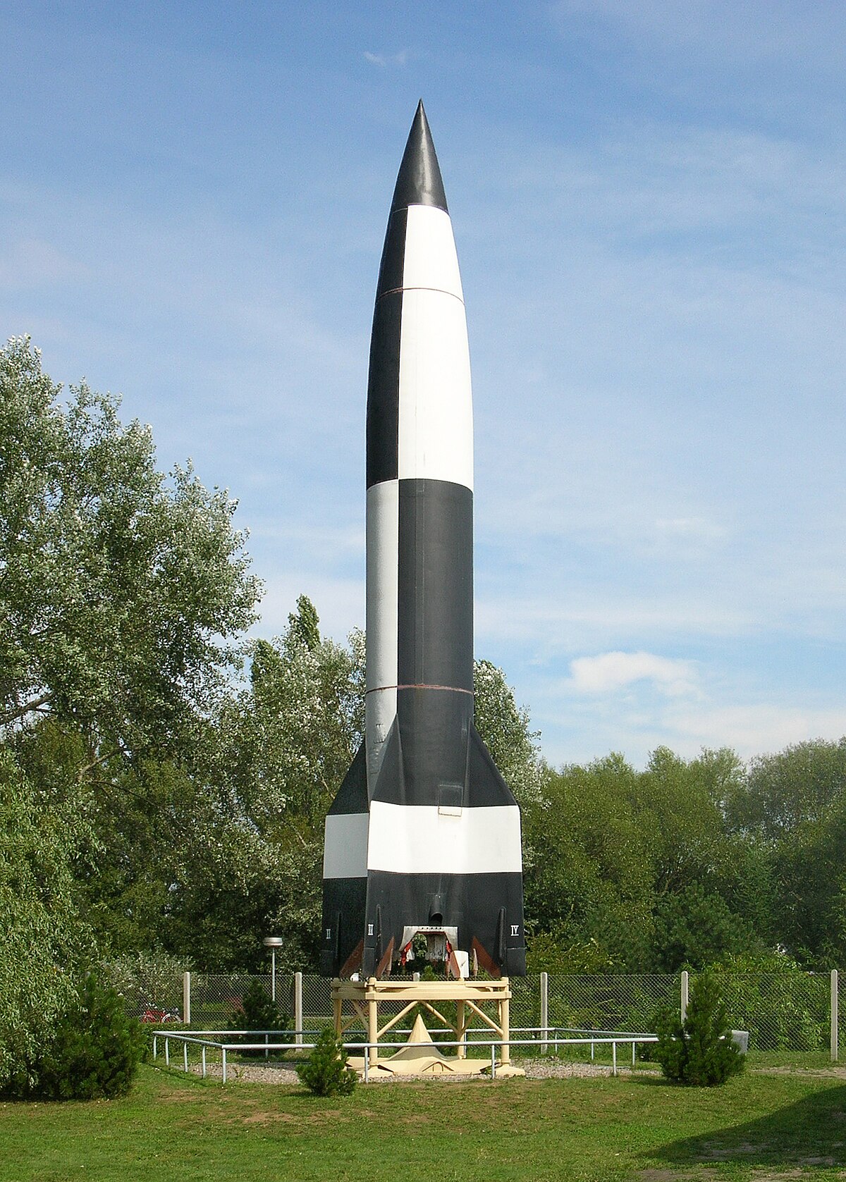 A V-2 Rocket, painted in a black and white checkered pattern, stands upright on a display stand at the Peenemünde Museum. The rocket is surrounded by green trees under a clear sky.