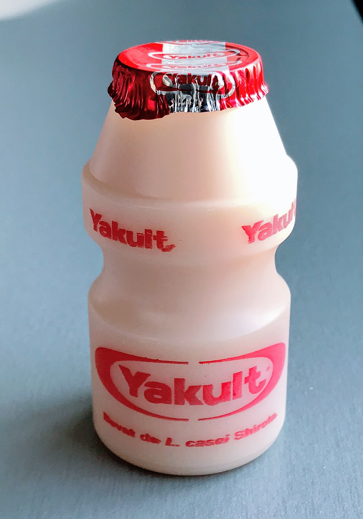 A small plastic bottle of Yakult, a probiotic drink, is shown with its red foil cap. The label on the bottle reads "Yakult" and "Bevat de L. casei Shirota" (Contains L. casei Shirota).