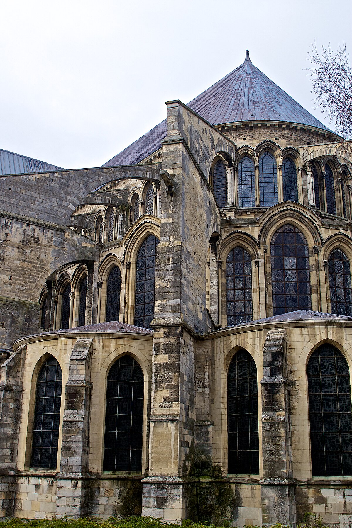The exterior of the Basilica of Saint-Remi in Reims, France, displays a series of flying buttresses and arched windows. The stone structure is weathered, and a conical roof is visible at the top.