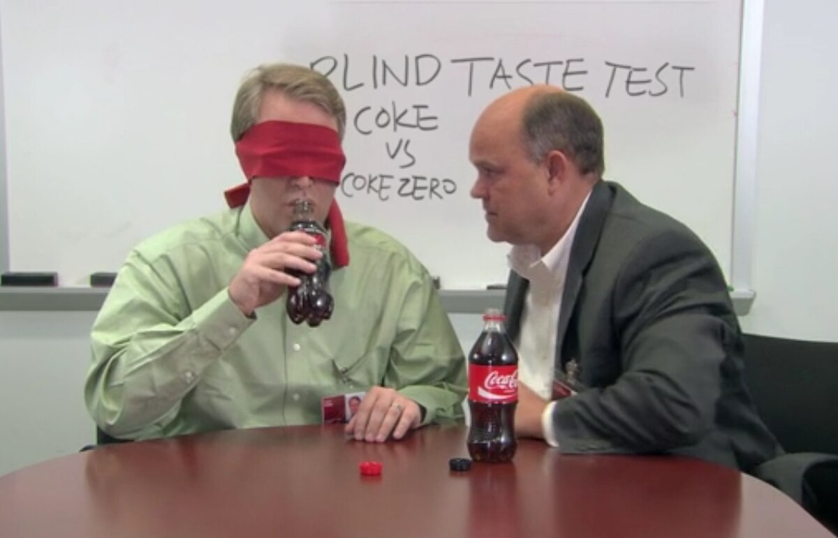 A single-blind taste test in progress, with a blindfolded man tasting a cola while another man observes. A whiteboard in the background reads "BLIND TASTE TEST COKE vs COKE ZERO".